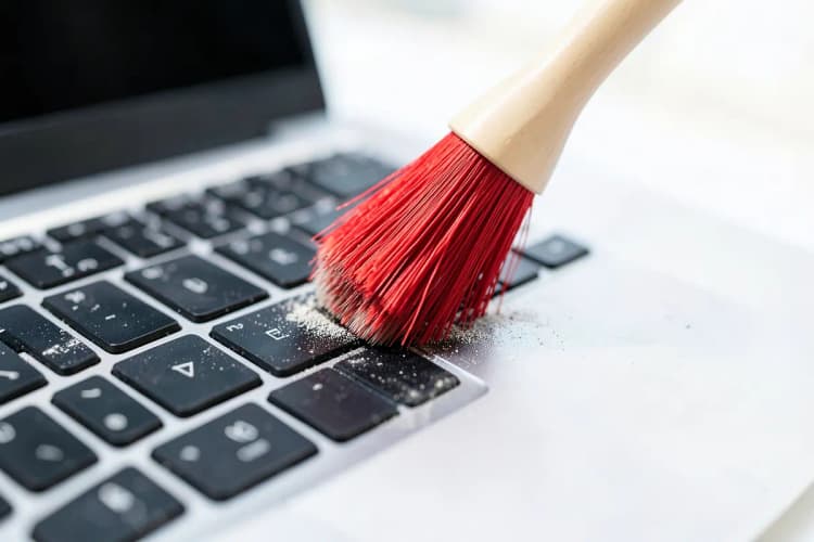 Uma mão segurando um pincel limpando a poeira de um teclado de notebook.